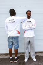Two models wearing white graphic long-sleeve t-shirts from the back and front, standing in front of a white corrugated metal wall.